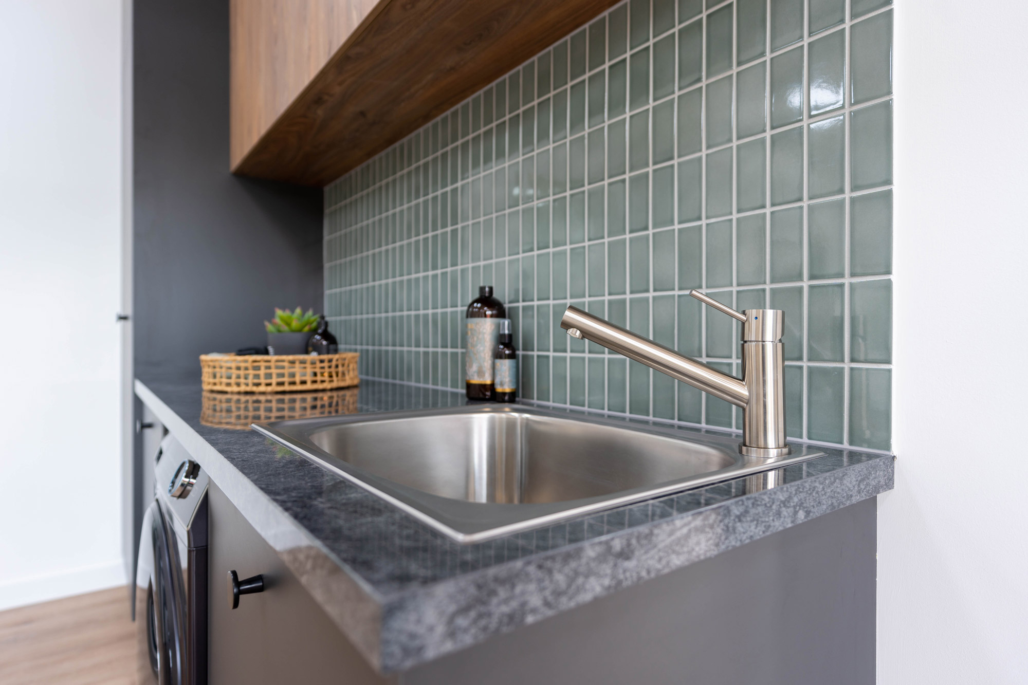 Close up photo of a laundry Oliveri sink and mixer tap. Sage green tile splashback with dark timber overhead cabinets