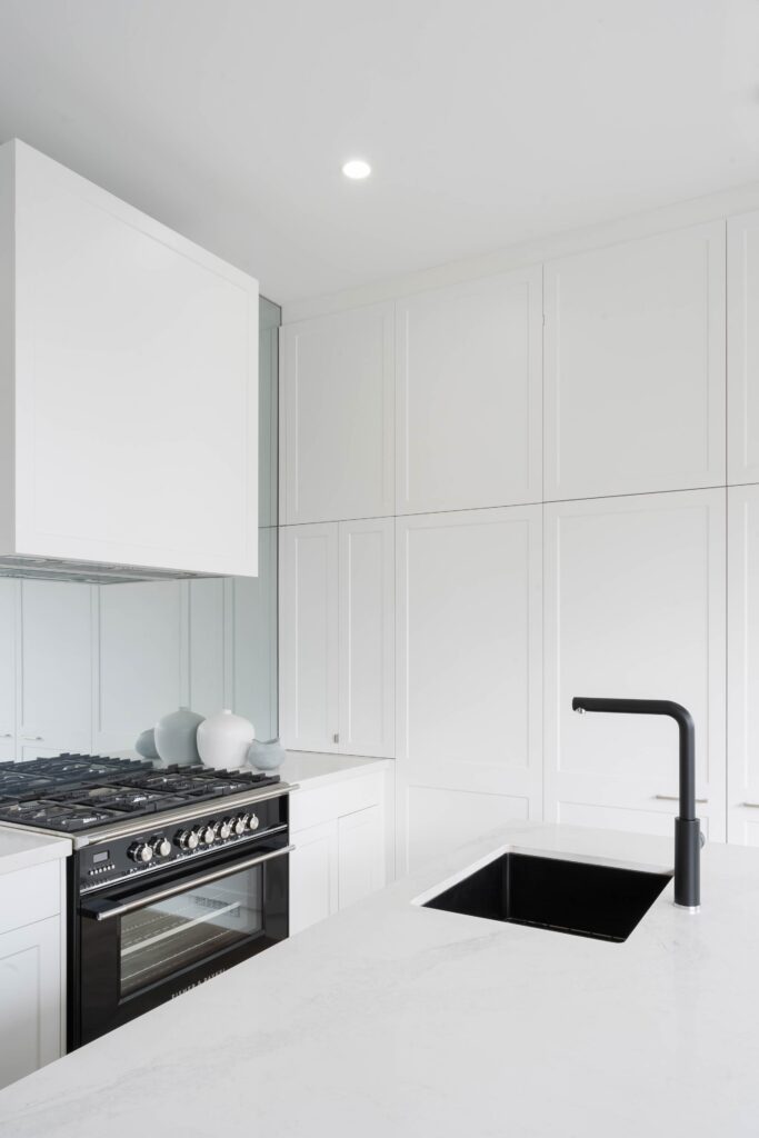 White Shaker style kitchen cabinetry with glass mirror splashback and a Fisher & Paykel freestanding black oven. White stone benchtops with black undermount sink and black mixer tap