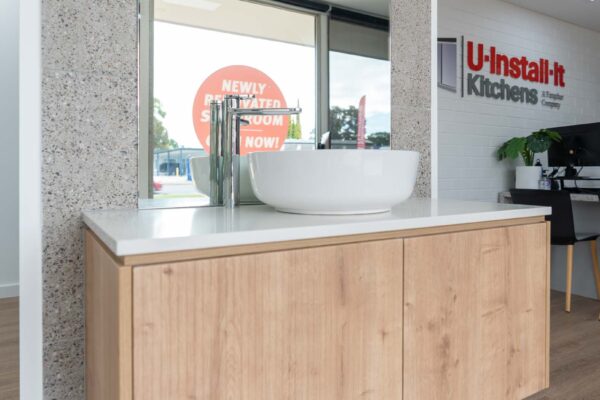Display room showing a bathroom vanity in a timber grain laminate with a stone white benchtop. Round white basin with an arched mirror and terrazzo tiles