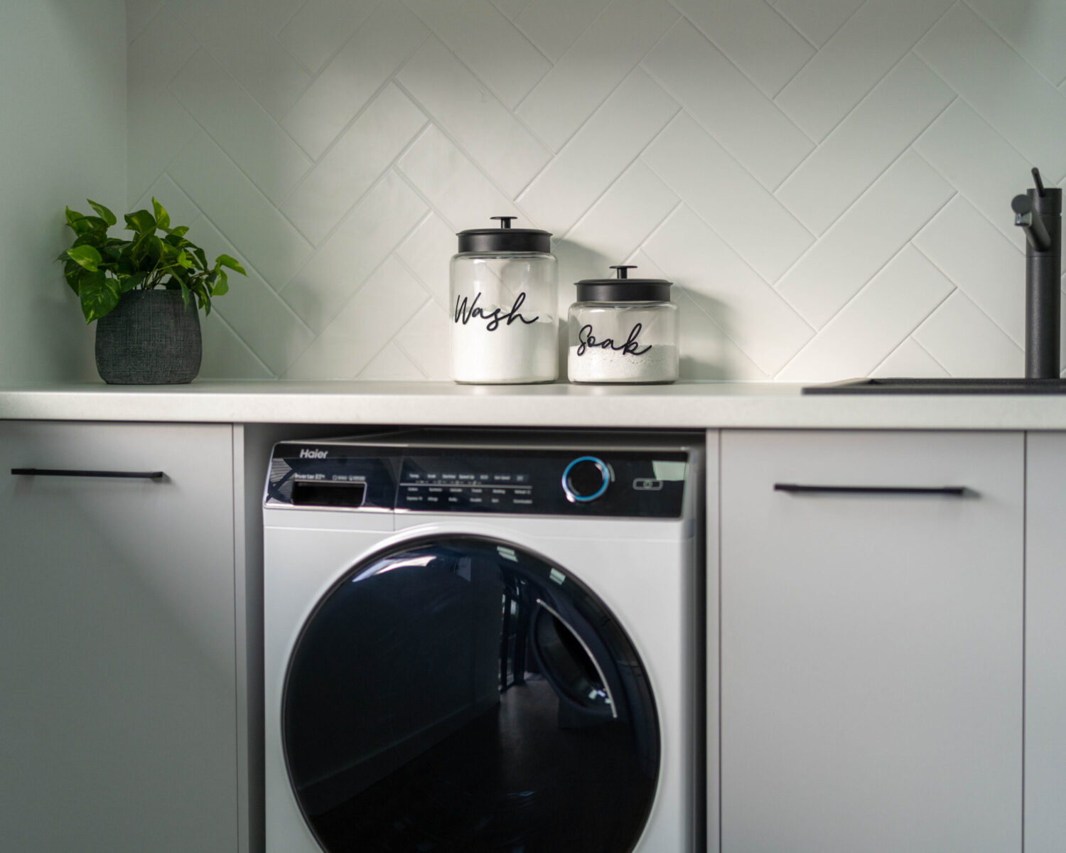 Laundry with white grey cabinetry, white laminate benchtop and white subway tiles in a herringbone pattern.