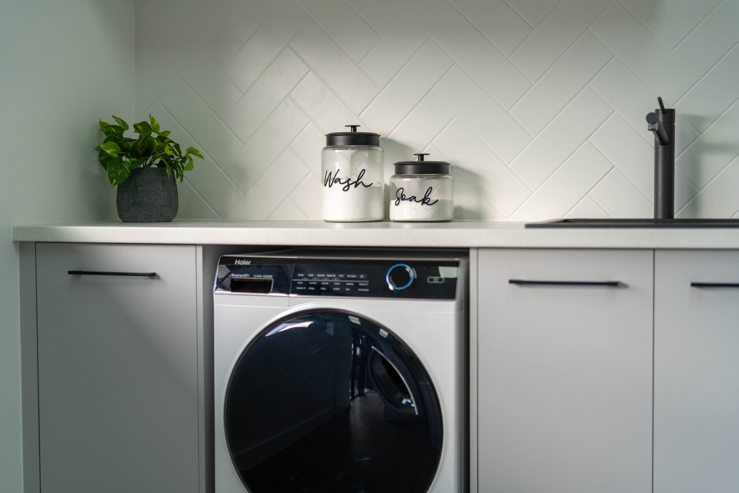 Laundry with white grey cabinetry, white laminate benchtop and white subway tiles in a herringbone pattern.